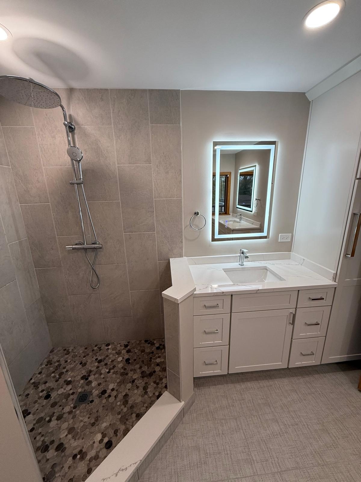 Walk-in shower with rain shower head and pebble floor next to a white vanity with LED mirror